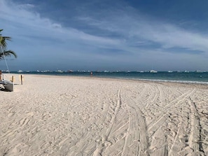 A sandy beach with tire tracks leading to the water, boats in the distance, and a clear sky. - Apartamento en Punta Cana para 4 personas (Punta Cana)