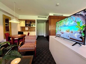 A hotel room with a dining area, a TV displaying a tropical beach scene, and a hallway leading to another room.