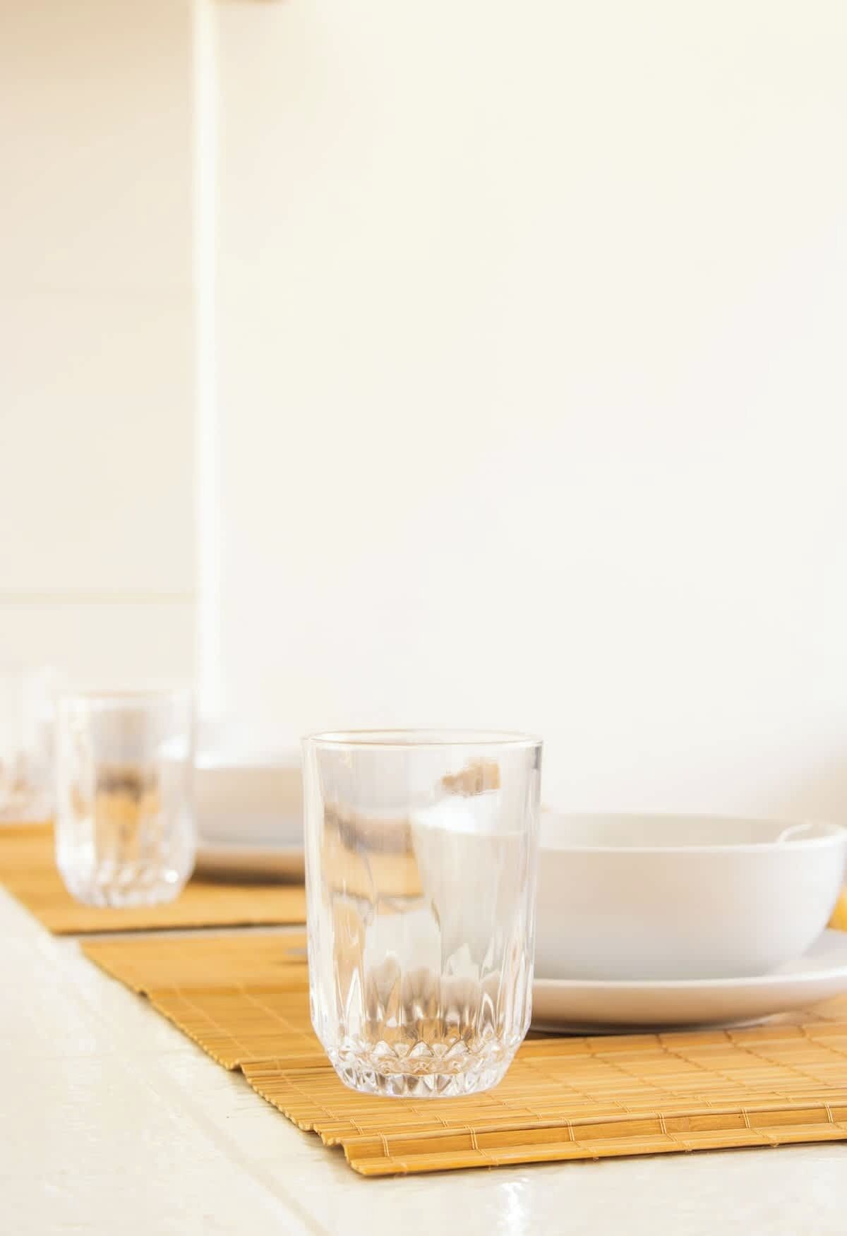A clear glass on a bamboo mat with a white plate and another glass in the background.