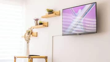 Een moderne eetruimte met een televisie aan de muur, een bamboe stoel, een eettafel met een kom en bestek, en een vaas met bloemen.