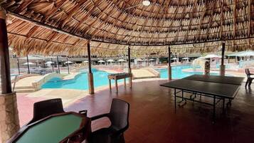 Un espace piscine extérieur doté d’un toit de chaume, de chaises longues et d’une table de ping-pong.