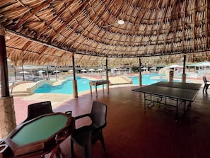 Un espace piscine extérieur doté d’un toit de chaume, de chaises longues et d’une table de ping-pong.