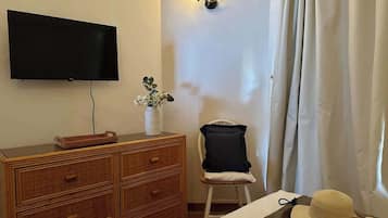 Une pièce avec un téléviseur à écran plat fixé au mur, une commode en osier, une petite chaise, un vase décoratif avec des fleurs et un chapeau posé sur le sol.