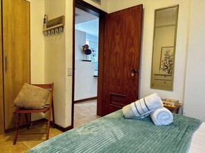 Habitación de hotel con cama, una silla, una puerta de madera, un espejo y un pasillo que lleva a un baño.
