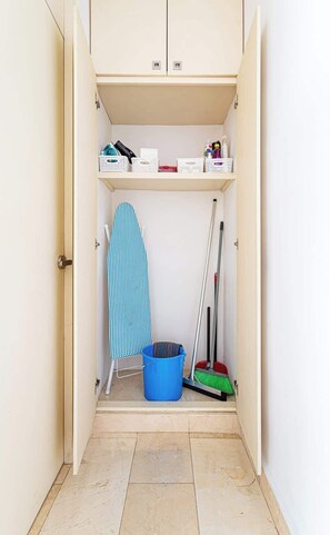 Ein Waschraum mit einem blauen Bügelbrett, einem Mop, einem Besen und Regalen.
