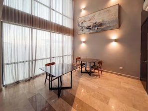 A dining area with a glass table and chairs, a painting on the wall, and a large window with curtains.