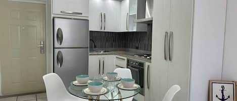 Une cuisine compacte avec des meubles blancs, un réfrigérateur en acier inoxydable, une cuisinière à gaz et une table à manger en verre, assortie de chaises blanches.