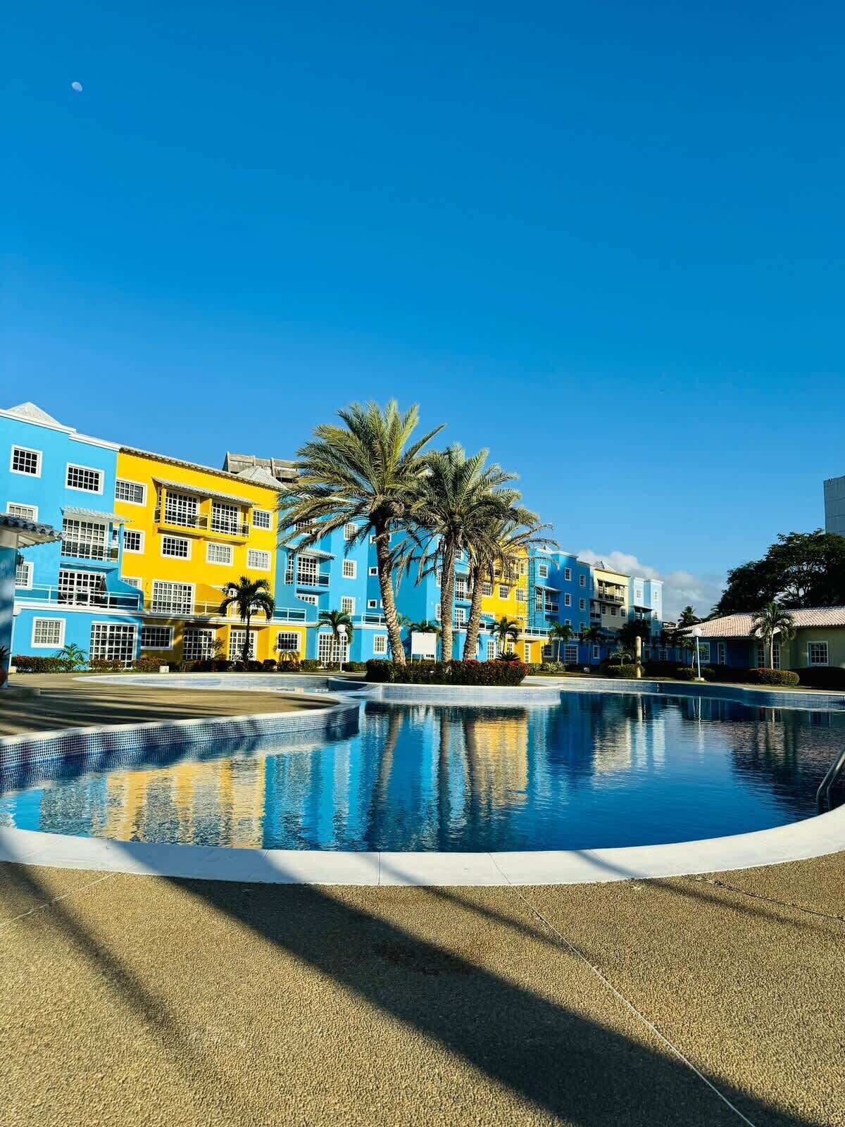 A swimming pool with a clear blue sky, surrounded by colorful buildings and palm trees.