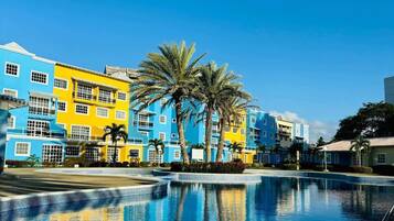 A swimming pool with a clear blue sky, surrounded by colorful buildings and palm trees.