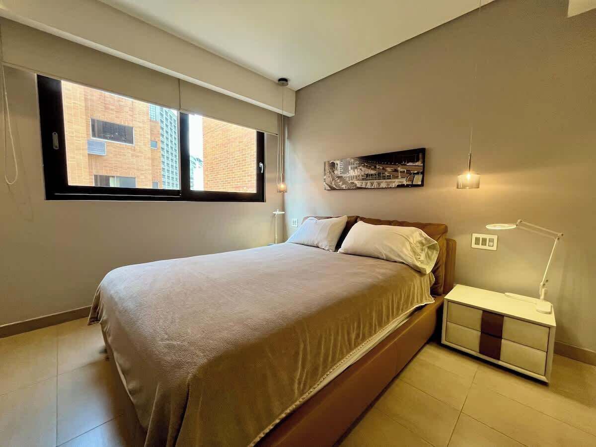 Een slaapkamer met een bed, nachtkastje, lamp en uitzicht op gebouwen door het raam.