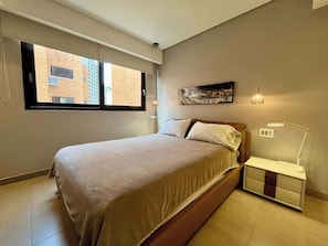 A bedroom with a bed, bedside table, lamp, and a view of buildings through the window. - Lux+comfort at Campo Alegre|Design & connectivity (Caracas)