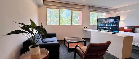 A hotel room with a bed, a desk with a computer, a chair, a small table with a plant, and a window.
