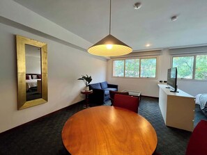 Ein modernes Hotelzimmer mit einem runden Holztisch, einer Couch, einem Fernseher und einem großen Fenster mit Blick auf Bäume.