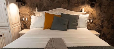 A bed with white linens, two pillows in different colors, and a stone wall behind it.