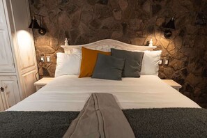 Una cama con sábanas blancas, dos almohadas de colores diferentes, y una pared de piedra al fondo.