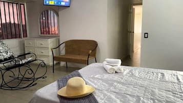 Hotellihuone, jossa on sänky, seinälle asennettu televisio, rottinkituoli ja ikkuna, jossa on sälekaihtimet.