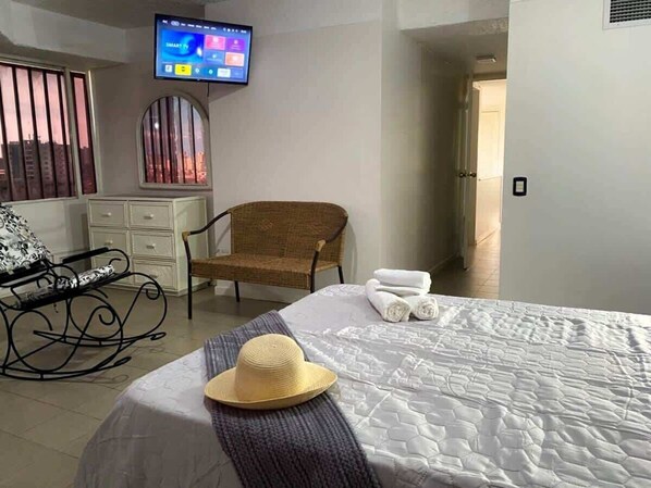 Une chambre d’hôtel avec un lit, un téléviseur fixé au mur, un fauteuil en osier et une fenêtre avec des stores.