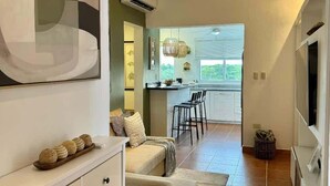 Uma sala de estar moderna com um sofá, uma área de jantar com cozinha ao fundo e uma grande janela que oferece luz natural.