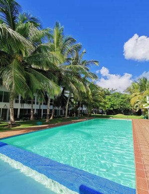 A swimming pool with clear blue water, surrounded by palm trees and a building with a white facade. - Apartment w/ balcony + Pool | Playa Bibijagua (Punta Cana)
