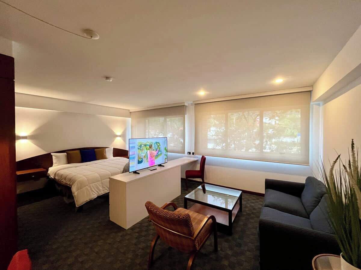 Habitación de hotel con cama, escritorio con televisor, sofá, sillón y ventana grande.