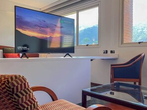 A living room with a flat-screen TV displaying a sunset, a glass coffee table, a patterned armchair, and a wooden chair. - Tu Rincón en Altamira: ¡Céntrico y Nuevo! (Caracas)