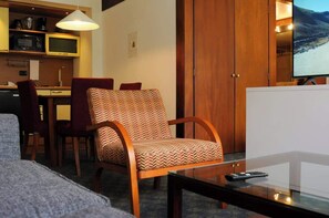 Een ruimtelijk ingerichte woonruimte met een bank, een gestreept fauteuil, een eethoek met stoelen, een televisie en een keuken met apparatuur.