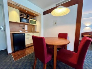 Een compacte keuken met een magnetron, koffiezetapparaat en een kleine koelkast. Er is een eethoek met een houten tafel en rode beklede stoelen.