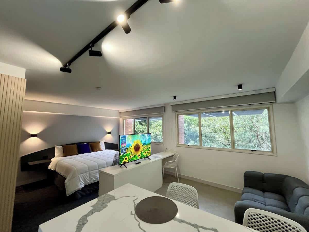 Una habitación de hotel moderna con una cama, un escritorio con computadora, un televisor y una ventana con vista a árboles.