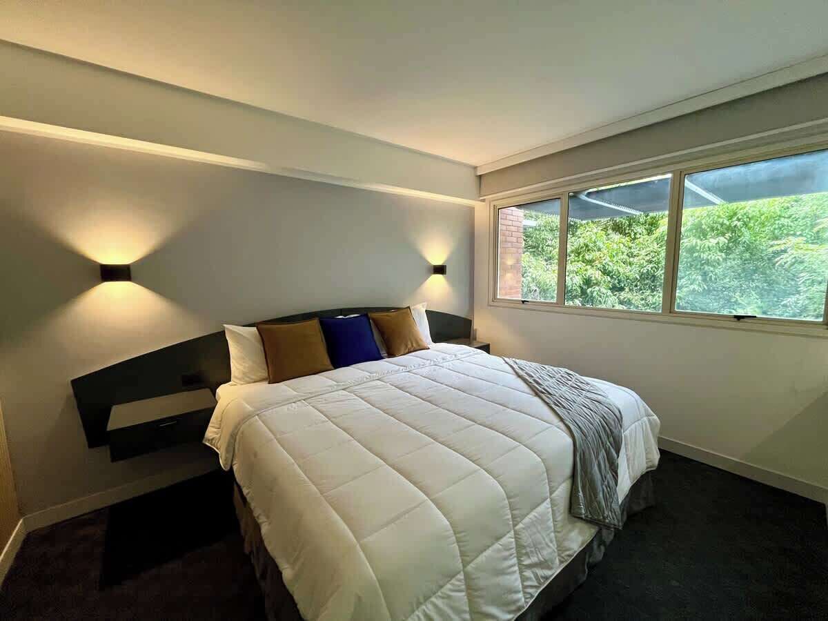 Un dormitorio con cama, mesita de noche, luces empotradas en la pared y ventana con vistas a zonas verdes.
