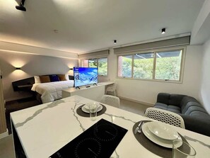 A modern hotel room with a dining area, a bed, a TV, and large windows.