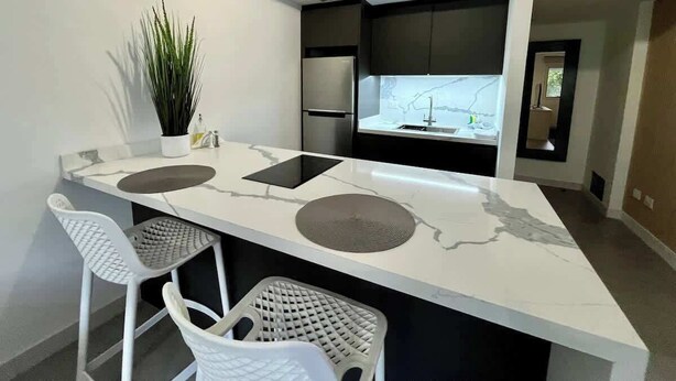 A modern kitchen with a central island, two bar stools, a potted plant, and a view of the living area. - Disfruta de esta acogedora habitación en Caracas (Caracas)