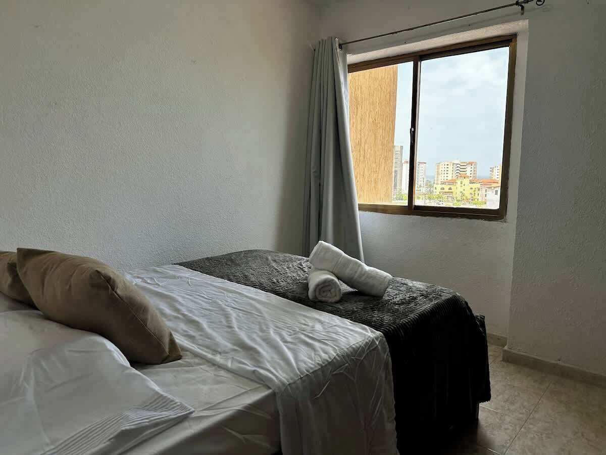 A room with a bed, a small table, a window with curtains, and a view of buildings outside.