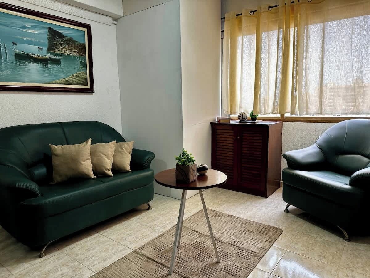 Ein Wohnzimmer mit einem grünen Sofa, einem schwarzen Sessel, einem kleinen runden Tisch mit einer Pflanze und einem Gemälde an der Wand.
