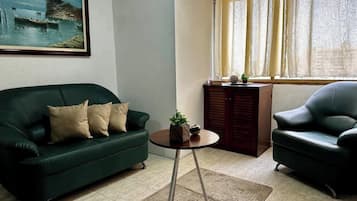 Ein Wohnzimmer mit einem grünen Sofa, einem schwarzen Sessel, einem kleinen runden Tisch mit einer Pflanze und einem Gemälde an der Wand.