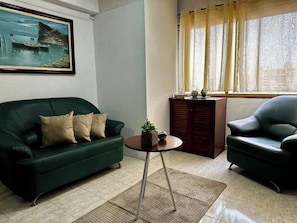 A living room with a green sofa, a black armchair, a small round table with a plant, and a painting on the wall.