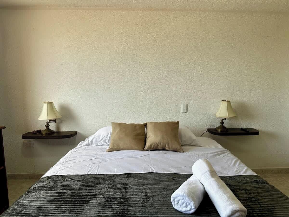 Ein Hotelzimmer mit einem Bett, zwei Nachttischlampen, einem zusammengerollten Handtuch und einer gemusterten Decke.