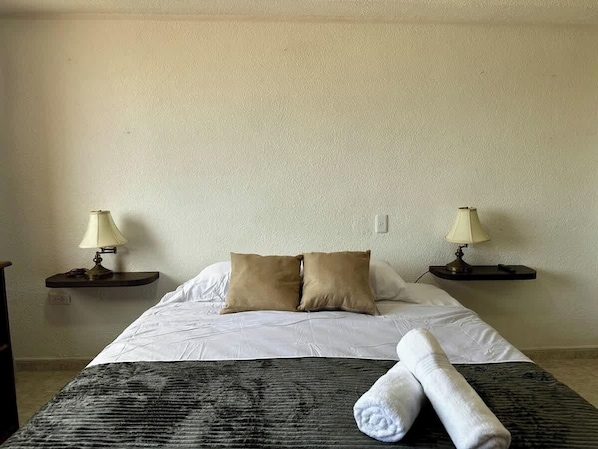 A hotel room with a bed, two bedside lamps, a rolled towel, and a textured blanket.