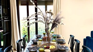 Un espace repas avec une longue table dressée pour six personnes, entourée de chaises, agrémenté d’un centre de table décoratif et offrant une vue sur la verdure à l’extérieur.