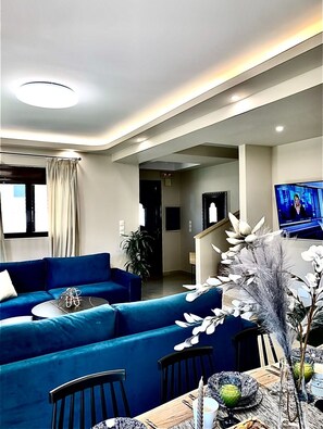 A modern living room with a blue sofa set, a dining table with chairs, and a television displaying a news program. - Hillside Cabana Maisonette in Koskinou Rhodes (Kallithea)