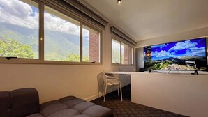 Una stanza con una grande finestra, una TV a schermo piatto, un divano e una scrivania con una sedia.
