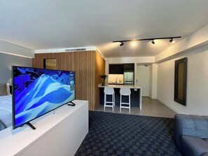 Ein modernes Hotelzimmer mit Fernseher, einer Sitzecke, einem Essbereich mit zwei Stühlen und einer Küche im Hintergrund.