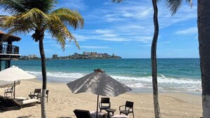 A beach with palm trees, a beach umbrella, and chairs. - Tu Refugio Porlamar | A pasos de Playa Veleros (Porlamar)