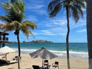 A beach with palm trees, a beach umbrella, and chairs. - Tu Refugio Porlamar | A pasos de Playa Veleros (Porlamar)