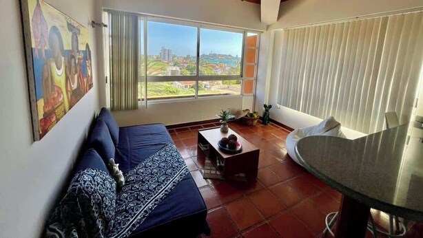 A living room with a sofa, a coffee table, a dining table, and a view of the cityscape through large windows. - Tu Refugio Porlamar | A pasos de Playa Veleros (Porlamar)