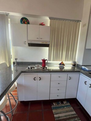 A kitchen with white cabinets, a stove, a red coffee pot, and a striped rug on the floor. - Tu Refugio Porlamar | A pasos de Playa Veleros (Porlamar)