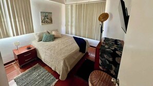 A bedroom with a bed, bedside table, lamp, and a window with blinds. - Tu Refugio Porlamar | A pasos de Playa Veleros (Porlamar)