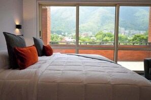 A bedroom with a large bed, two pillows, a nightstand, and a window with a view of a mountainous landscape. - PH Altamira: Vistas Ávila & Terraza Panorámica (Caracas)