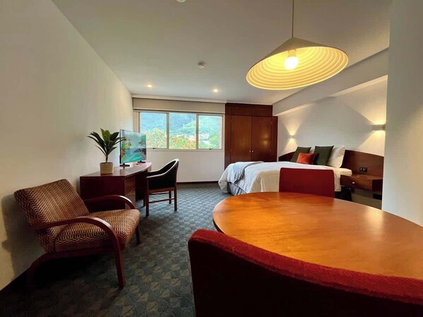 A hotel room with a bed, a desk, a chair, a dining table, and a large window.