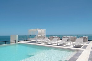 Ein Infinity-Pool auf dem Dach mit Liegestühlen und einer Glasreling mit Blick auf den Ozean.