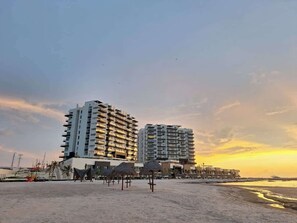 Ein Hotel direkt am Strand mit mehreren Etagen, ein sandiger Strand im Vordergrund und ein klarer Himmel.
